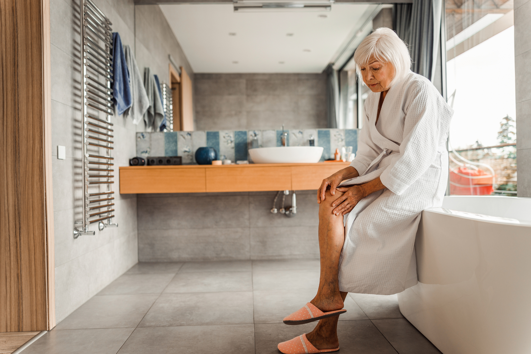Older adult sitting on the edge of a traditional tall bathtub wall, illustrating the fall risk of standard bathroom design for seniors.
