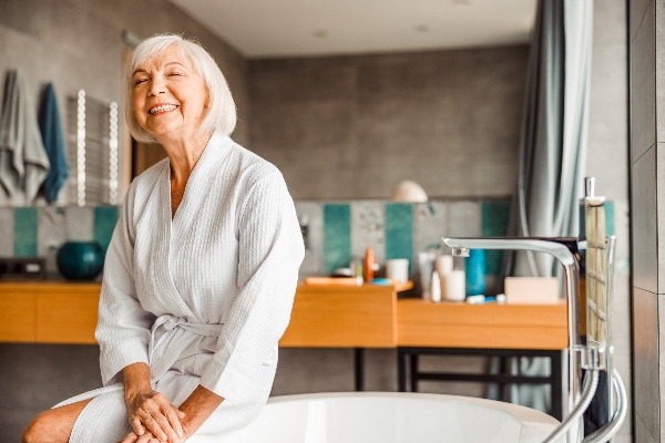 A senior smiles next to a walk-in bathtub, something that has many benefits for her.